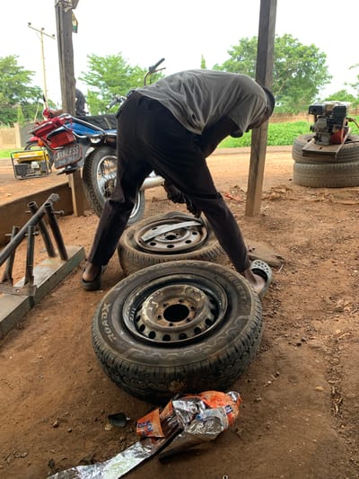 In Gbeleko, Nigeria, a man wearing dark pants and a grey t-shirt is bent over a tire on a dirt ground. He is wearing black clogs. There are several tires stacked nearby, and a generator is on top of two tires in the background. A motorcycle with a blue fuel tank and a red rack is parked behind him. The scene appears to be an outdoor workshop or repair area.