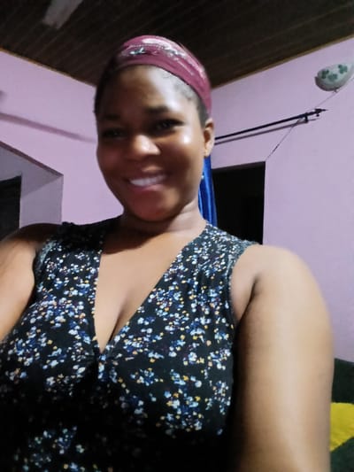 The image shows a medium shot of a woman with dark skin, smiling and looking at the camera. She is wearing a dark floral print top with a V-neck and a maroon headscarf. Her hair is not visible. The background is a room with light purple walls. A dark ceiling with wooden panels is visible at the top. A black cable runs across the wall. On the right side of the wall, there is a white object with green markings, possibly a decorative item. To the right of the woman, a dark doorway is visible. The lighting appears to be artificial, casting a slight sheen on her skin. The scene is indoors and does not provide any specific visual cues to pinpoint its location within Yenegoa, Nigeria, beyond it being an interior residential or personal space.