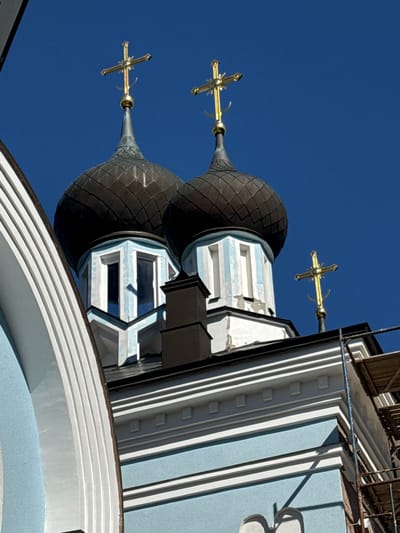 The image shows the exterior of an Orthodox church in St. Ozerki, Russia. The focal point is the upper section of the building, featuring multiple onion domes. Two prominent domes, covered in dark, segmented material, are visible, each topped with a golden cross. The domes are supported by white, multi-faceted structures with blue accents and windows. A third, smaller golden cross is visible to the right, also atop a dome or spire. The sky is a clear, deep blue, contrasting with the dark domes and the light blue and white facade of the church. The lower portion of the image reveals architectural details of the building, including decorative white horizontal banding and some scaffolding on the right side, suggesting ongoing construction or maintenance.