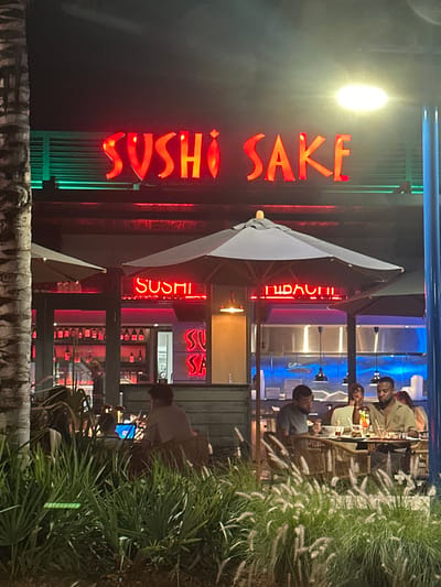 The image displays the exterior and outdoor dining area of the "Sushi Sake" restaurant at night in Lauderdale-by-the-Sea, United States of America. The restaurant's name, "SUSHI SAKE," is prominently displayed in large red neon letters across the upper facade of the building. Smaller red neon signs reading "SUSHI" and "HIBACHI" are visible below the main sign, within the restaurant's windowed section.
The foreground features dense green and white ornamental grasses, with a palm tree trunk partially visible on the far left. The outdoor dining area includes multiple round tables with wicker chairs, several of which are covered by large grey patio umbrellas.
Numerous individuals are present. On the left side of the outdoor seating, one person is seen from the back, seated at a table. On the right, four individuals occupy another table; three are clearly discernible. One male with a beard, wearing a light-colored polo shirt, is seated on the far right, looking down at the table. To his left, another male with a beard, wearing a grey t-shirt, is looking left. Between them, a female in a light jacket or blazer is seated, facing right. Glasses containing beverages and plates are visible on their table. Inside the restaurant, behind large glass windows, a bar area stocked with bottles is visible on the left, where a person is standing. Further right inside, a brightly lit area, possibly a kitchen or prep station, features blue accent lighting. The scene is illuminated by the restaurant's signage, streetlights, and internal lighting.