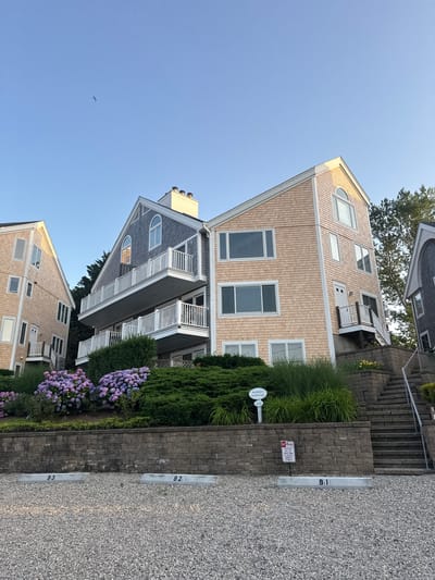 In Provincetown, Massachusetts, USA, a multi-story residential building with light peach-colored shingle siding is photographed at a low angle, showcasing its distinctive architecture. The building features multiple balconies with white railings and several windows, including a prominent arched window at the top. Adjacent to it is a building with gray shingle siding. The foreground is a gravel parking lot with concrete parking space markers labeled B1, B2, and B3. Behind the parking lot, a low stone wall supports a terraced garden area with lush green bushes and vibrant purple hydrangeas. A sign on a post in the garden reads "BUILDING B 26 WEST VINE." A red and white "NO PARKING" sign is also visible on the stone wall, indicating that violators will be towed. The sky above is clear and blue, with a single bird in flight. The scene is illuminated by natural light, suggesting it is either early morning or late afternoon.