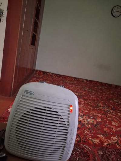 The image shows a close-up of a white electric fan heater with a red indicator light on. The fan is positioned in front of a red carpet with a floral pattern. To the left of the fan, a light-colored wall is visible, and further left, a clock with a white face and black numbers is mounted on the wall. The clock shows the time as approximately 12:09. Below the wall and clock, there is a wooden surface that appears to be part of a piece of furniture or a floor. The overall scene suggests an indoor setting, possibly a room in a home.
Visible text on the fan includes "De'Longhi" and "VERTICALEFYOUNG". The fan appears to be operating, indicated by the lit red light. No people are visible in the image. The time of day is indicated by the clock, and there are no clues about the weather or specific emotions. The location context provided is Kahramon, Uzbekistan.