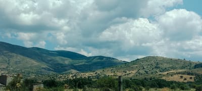 The image depicts a landscape scene with rolling hills and mountains under a cloudy sky. In the foreground, there are remnants of abandoned structures, appearing to be old buildings with exposed brickwork and weathered surfaces. The terrain is covered with vegetation, including sparse trees and dry grasses on the hillsides. The mountains in the background are covered in a mix of greenery and brown, indicating arid or semi-arid conditions. The sky is filled with a mixture of light and dark clouds, suggesting either a partly cloudy or overcast day. The scene appears to be situated in a rural or mountainous area, possibly in the vicinity of El Espinar, Spain, given the rocky and scrubland vegetation. No people or animals are visible in the image.