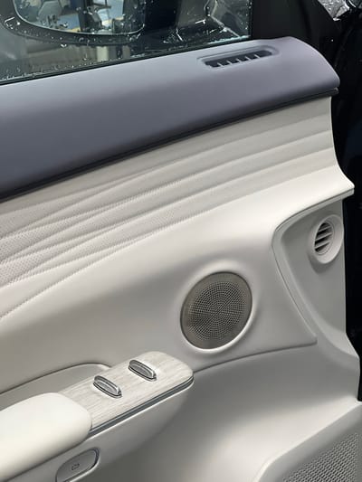The image shows the interior of a car door. The door panel is predominantly white with a textured, perforated insert. A speaker is visible on the lower portion of the panel. Above the speaker is a circular air vent. The armrest has a wood-grain accent with two silver buttons and another silver button located lower down. The top of the door panel is a dark grey material with a horizontal air vent. The window reflects an exterior scene with water droplets. The scene does not provide any information about Kabanikha, Russia.