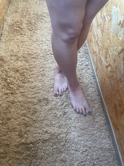 The image shows a close-up of a person's legs and feet standing on a beige shag carpet. The person's feet are crossed, and their toenails are painted dark gray. The background consists of a wall made of oriented strand board (OSB). The scene appears to be indoors, possibly in a room with unfinished walls. There is no indication of the city of Myrhorod, Ukraine, in the image.