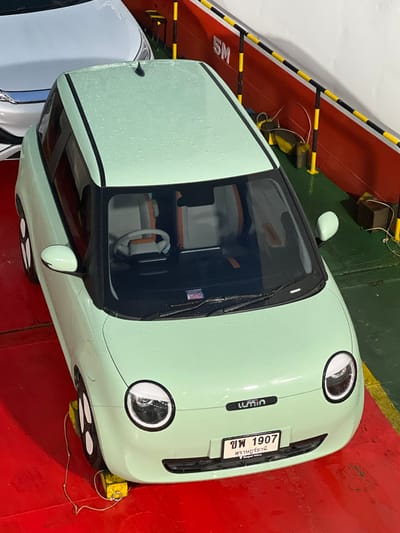 A light green, compact car, possibly an electric vehicle, is parked on a red, metal surface, likely a ferry deck. The car's surface is speckled with water droplets, suggesting it might be raining or has recently rained. The car has a black trim around the windows and a black stripe along the side. Its front features two large, round headlights and a license plate.
The license plate is white with black text, reading "ขพ 1907" at the top. Below this, in smaller text, are Thai characters and "Deepal Wavy". Another car, light blue or white, is partially visible to the left of the green car.
The scene is bordered by yellow and black striped safety barriers. In the background, there are wooden blocks and ropes. The overall impression is one of a functional, utilitarian setting for transporting vehicles, possibly a ferry or a parking deck on a ship. The lighting appears to be artificial, consistent with an indoor or covered space. There are no people visible in the image. The time of day and specific location are not discernible from the visual cues, beyond the general context of vehicle transport.