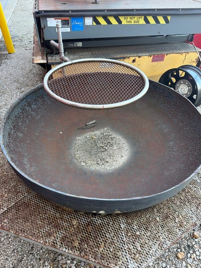 This is an outdoor, eye-level, medium shot photograph of a large, rusted metal fire pit. The fire pit is round and sits on a metal grate. In the background, there is a yellow lift gate with a "DO NOT LIFT THIS SIDE" sticker. A portion of a black tire rim is visible to the right of the fire pit. The ground surrounding the objects is covered in gravel. The scene appears to be in Hornersville, United States, though no specific landmarks are visible.
