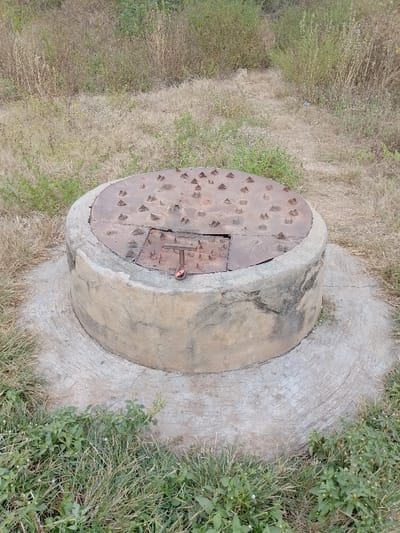 A circular, concrete structure with a rusty metal lid sits amidst sparse vegetation. The lid is studded with pyramid-shaped spikes and has a small, hinged opening with a T-shaped handle, where a pink object hangs. The structure rests on a circular concrete base. The surrounding area is a mix of dry grass and patches of green vegetation.