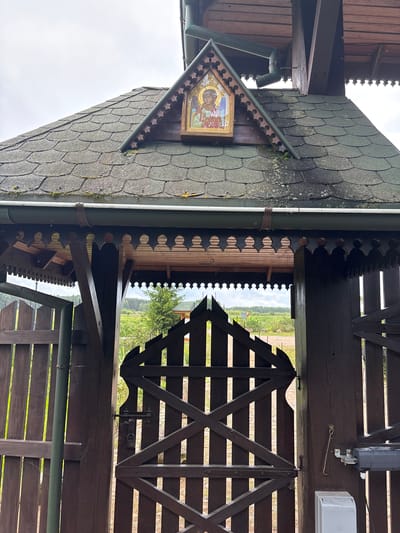 An Orthodox icon of an angel is affixed to a triangular wooden gable above a weathered, dark brown wooden gate in a rural setting in Koleśne, Poland. The roof over the gate is covered in dark green shingles. A rain gutter runs horizontally across the top of the gate. The gate itself is constructed of dark wooden planks and features an ornate, cutout wooden trim along the top. The gate is secured with a metal latch and a padlock. Beyond the gate, a pastoral landscape with trees and fields is visible under a cloudy sky. The ground in front of the gate appears to be gravel or dirt. A portion of a dark brown wooden fence is visible to the left of the gate.