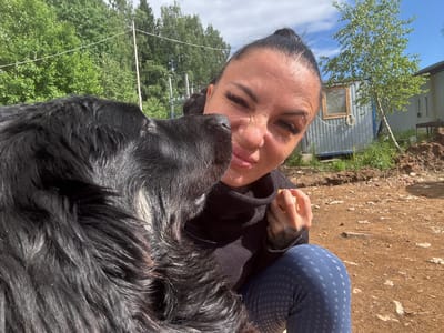 In Dulepovo, Russia, a woman with dark hair pulled into a bun and wearing a black turtleneck and blue patterned leggings is being nuzzled by a large black dog in a close-up selfie. The woman's eyes are closed and she is smiling, while the dog's nose is pressed against her cheek. Her left hand is raised and holding a piece of food near her face, and she has a tattoo on her forearm. The background shows a rural outdoor setting with green trees, a corrugated metal building with a window, and an area of disturbed earth. The ground is covered in dirt, small rocks, and patches of dry grass. The sky is blue with scattered white clouds.