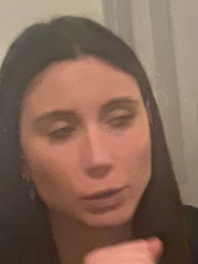 A blurry, close-up, head-and-shoulders portrait depicts a female subject with dark hair and fair skin. Her head is angled slightly to the right, with her gaze directed downwards and her eyelids partially closed. Her mouth is slightly agape. A small, dark, possibly dangling earring is visible on her left earlobe. The fingers of what appears to be her right hand are partially visible in the lower right quadrant, obscuring part of her chin and lower lip. The background is indistinct, appearing as a light-colored, plain interior wall or surface with a faint vertical texture or pattern on the right side. The image quality is low, characterized by significant blur and lack of sharp detail. The specific location within the city of Roanne, France, is not visually discernible from the image content.
