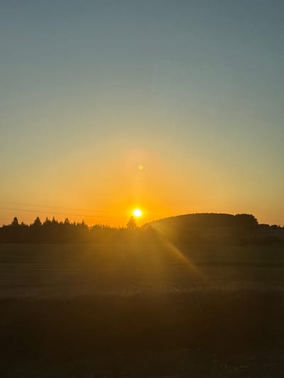 The image shows a landscape at sunset, potentially near Bol'shiye Klyuchishchi, Russia. The sun is setting on the horizon, visible slightly above a line of trees and casting a bright yellow and orange glow. A lens flare is also visible. The sky is a gradient, fading from blue on the left to orange near the horizon. To the right of the sun, there's dark vegetation that might be a field. The photo appears to be taken from a slightly elevated perspective, maybe from a field. The image is rotated to the side, making the horizon appear vertical.