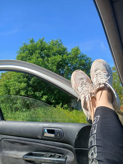 The photograph depicts a person's legs resting on the open door of a car, presumably while parked in a natural setting. The person is wearing black leggings with white text on the lower leg and light-colored sneakers. The legs extend from inside the car, resting on the upper door frame. The car's interior door panel is visible with a handle and window controls. Outside the car, there is a lush green tree against a clear blue sky. The location is claimed to be Sokolovo, Bulgaria. However, there are no landmarks or indicators to definitively confirm this geographic location. The composition is rotated.