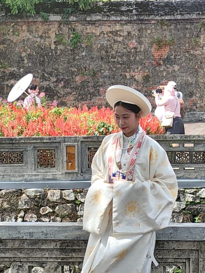 The scene is set outdoors in Phú Xuân Ward, Huế, Vietnam, likely within a historical complex such as the Imperial City. In the foreground, a young East Asian female stands centrally, facing slightly to her left. She wears a wide-brimmed, flat white traditional Vietnamese hat and an elaborate cream-colored long-sleeved robe with gold floral patterns and a complex multicolored embroidered collar. A multi-strand pearl necklace is draped around her neck. Her hands are clasped or folded at her waist, and her gaze is directed downward.

Behind her, a grey stone balustrade with ornate square cutouts extends horizontally. Beyond this balustrade, a dense bed of red flowering plants forms a vibrant mid-ground. To the left, partially obscured by these flowers, a second female in light-colored attire holds an open white parasol. To the right, a third female, dressed in a pink top, dark pants, and a white cap, is actively holding a smartphone or camera at eye level, appearing to photograph something beyond the frame. A fourth individual is partially visible behind her. The background consists of a tall, weathered brick wall, exhibiting discoloration and sparse green vegetation. Below the balustrade, a rough stone wall foundation is visible. The scene is brightly lit under direct daylight.