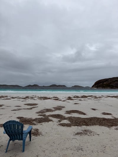 The image displays a coastal scene within Cape Le Grand, Australia. The sky is completely overcast with dense, grey clouds. In the foreground, a white sand beach is visible, extensively covered with irregular patches of dark brown marine vegetation. A single, unoccupied dark teal plastic Adirondack-style chair is positioned in the lower left quadrant of the image, facing towards the water. Beyond the beach, turquoise ocean waves break, leading to darker blue waters. A series of dark, rounded landmasses, consistent with islands or headlands, are visible along the horizon line. No individuals or active subjects are present in the scene.