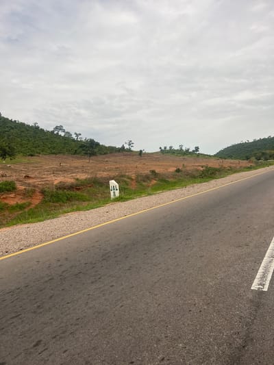 The image shows a roadside scene in Minda, Nigeria. The focus is on a highway with a gray asphalt surface and a yellow line marking the edge of the lane. A white concrete marker with green lettering, "JAL 8", is visible on the grassy verge beside the road. The land slopes upwards from the road, covered in dry earth, sparse green grass, and scattered trees. Further up the slope, the vegetation becomes denser, forming a green hillside. The sky is overcast with white and gray clouds, suggesting it is daytime. The overall impression is of a rural or semi-rural highway environment under cloudy skies. There are no people or vehicles visible in the image, and the scene appears quiet and still.