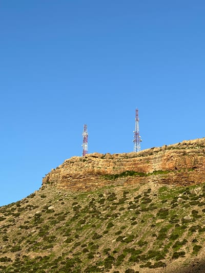 Two communication towers stand against a clear blue sky, positioned near the edge of a rocky cliff face. The cliff is covered with sparse green vegetation and exhibits distinct layers of brown rock. The towers are painted white and red, designed with a lattice-like structure. The scene is captured from a perspective looking upwards, emphasizing the height of the towers and the steepness of the cliff.