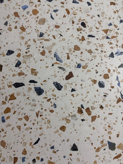 This is a close-up, top-down view of a terrazzo surface. The surface is a speckled off-white background with a variety of small, irregular chips of different colors embedded within it. The chips are predominantly shades of brown, tan, black, and dark blue. Some of the chips are quite small, almost like grains of sand, while others are larger, irregularly shaped fragments, some measuring over a centimeter in length. The texture of the background appears slightly rough and granular. There are no people, objects, or any discernible scene or setting visible. The image focuses solely on the pattern and texture of the terrazzo material itself. The lighting is even, suggesting an indoor environment with artificial light, or an overcast day. There is no visible text. The location context provided is Skopje, North Macedonia, but there are no visual cues within the image to support or contradict this information.