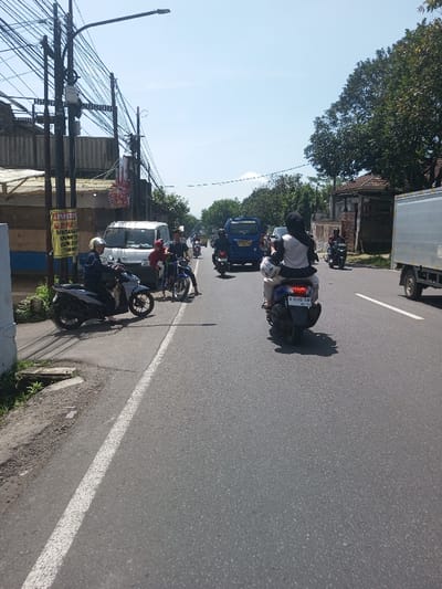 The image depicts a daytime scene on a paved two-lane road in Beteng_Pangkhar_Somthrang, Bhutan, under a clear, bright blue sky.
In the foreground on the left, a light-colored scooter is parked on a paved shoulder, with a male wearing a white helmet and dark attire seated on it, facing right. Adjacent to this, multiple utility poles heavily strung with electrical and communication wires stand, one bearing a yellow and white sign with dark text.
Midground, three individuals are gathered around a dark-colored motorcycle near the left edge of the main road. One person in a red top leans over the motorcycle, another in a dark shirt stands upright beside it. A white van is parked nearby, and a blue van with license plate "B 1587 FPJ" is visible behind it, facing away from the camera. Further right on the road, a dark blue scooter with license plate "B 5202 TAF" carries two individuals, the passenger wearing a white top and black hijab, both facing away from the camera. Other motorcycles and vehicles are visible further down the road, receding into the distance.
On the far right midground, a light grey truck is partially visible, also moving away from the viewer. The roadside is lined with trees and what appear to be residential or commercial structures. The background shows a continuation of the road, more trees, and faint hills or mountains on the horizon. Shadows are distinctly cast on the road, indicating bright sunlight.