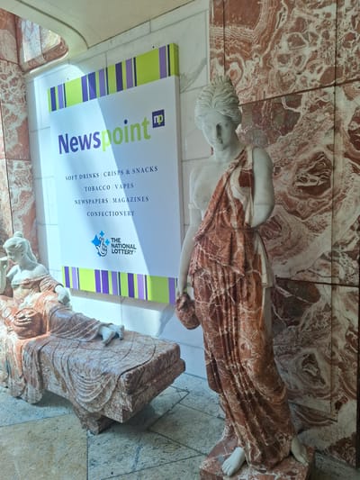 The image shows a Newspoint store with marble statues in front of it. The store has a sign that reads "Newspoint" with a logo next to it. Below the "Newspoint" text, it lists items for sale: "SOFT DRINKS CRISPS & SNACKS", "TOBACCO VAPES", "NEWSPAPERS MAGAZINES", and "CONFECTIONERY". Below this is the logo for "THE NATIONAL LOTTERY". The store is decorated with colorful stripes. The statues are of women in classical attire, made of what appears to be marble. One statue is reclining on a chaise lounge, while another is standing. The background is a wall of red and white marble. The floor is tiled. The scene is brightly lit, possibly by natural light coming from an overhead source. The location is stated to be Trafford, United Kingdom.