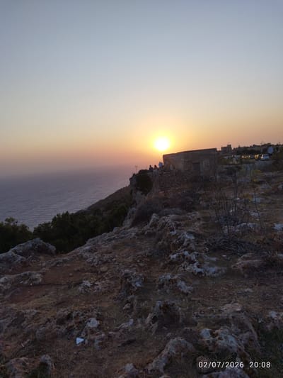 The image captures a sunset view from Dingli Cliffs, Malta. A vast expanse of the Mediterranean Sea stretches to the horizon, meeting a gradient sky of pale blue to soft orange. The rugged, rocky terrain of the cliffs dominates the foreground, sparsely covered with dry vegetation and small stones. On the cliff edge, a few people are visible, some seated and others standing, observing the sunset. Behind them, a weathered, stone building with a simple, unadorned facade stands prominently. Further in the background, several vehicles are parked, suggesting a popular viewpoint. The overall scene is tranquil, bathed in the warm, fading light of the setting sun.