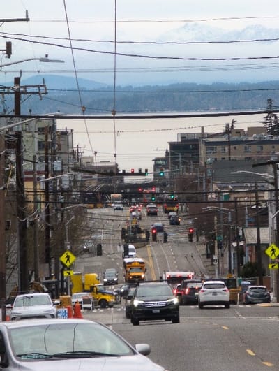 An elevated view showcases a bustling city street. Vehicles, including cars, vans, and a school bus, navigate the road. Traffic lights are visible, along with a pedestrian crossing sign. Overhead power lines crisscross the scene, framing the urban landscape. In the distance, a body of water is visible, with a cloudy sky above. Buildings of varying heights line the street, adding to the city's dense environment.