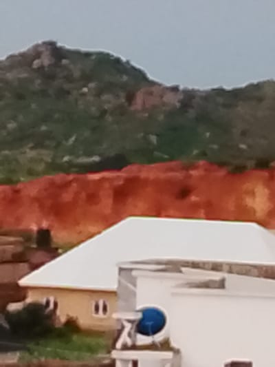 The image captures a view of a residential area in Gero, Nigeria, with a focus on buildings in the foreground and natural landscape in the background. The foreground features a light-colored, multi-story building with a prominent white roof. Adjacent to it, another building is partially visible, appearing to be a light yellow color with two windows. A blue satellite dish is mounted on the side of the white-roofed structure. Vegetation, appearing as green grass and bushes, surrounds the buildings. In the mid-ground, a large expanse of reddish-brown soil or rock formation is visible. The background is dominated by a green, rocky hillside or mountain. The sky above is a muted gray, suggesting overcast conditions or a hazy atmosphere. The image appears to be taken from a distance, with a somewhat low resolution. There are no discernible individuals or actions taking place in the scene.