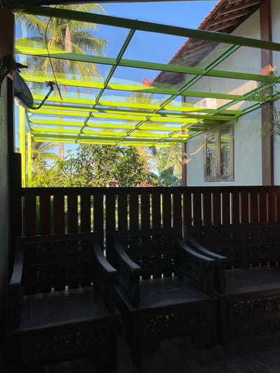 The image depicts a covered outdoor seating area with a green metal frame and glass roof. Three intricately carved wooden chairs are positioned in the foreground, casting shadows on the tiled floor. Beyond the chairs, a wooden fence separates the seating area from lush green foliage, including palm trees and other tropical vegetation, visible under a bright blue sky. A white building with a red-tiled roof and a wooden-framed window is visible to the right, with an air conditioning unit mounted on the exterior wall. The scene appears to be located in Kemloko, Indonesia, suggesting a tropical or subtropical climate. There are no people or animals visible in the image.