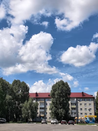 The image shows a residential building with a red roof and a light-colored facade. It is a multi-story building with many windows and balconies. In front of the building, there is a parking lot with several cars. Some trees are visible on the left side of the building. The sky is blue with white clouds. On the right side of the building, there is a small playground and a yellow sign that says "Меридиан-тур" (Meridian-tour). The scene appears to be a typical residential area in a Russian city. The image is taken from a slightly low angle, emphasizing the building and the sky.