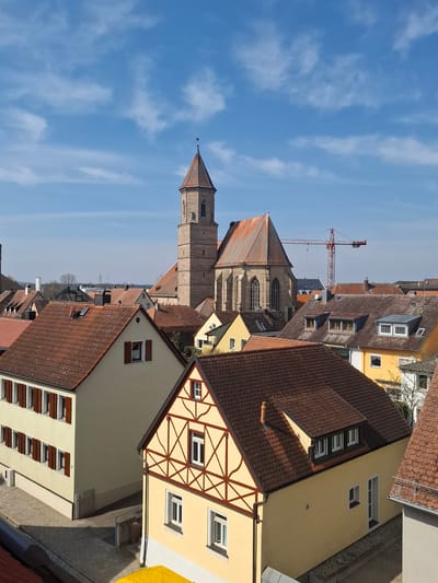 An elevated, clear-day view depicts a dense collection of buildings in Gunzenhausen, Germany, under a bright blue sky with scattered white clouds. The scene is devoid of visible people or active vehicles.
In the foreground, a light beige residential building with a reddish-brown tiled gable roof occupies the left side. It features multiple rectangular windows with closed dark brown wooden shutters on its two visible stories, and a white brick-patterned base. Adjacent to it, a light yellow half-timbered house with dark brown wooden beams arranged in cross and diagonal patterns stands prominently on the right, also with a reddish-brown tiled gable roof. This house displays white-framed windows, some with dark brown shutters, and a dark brown downspout. A paved sidewalk runs between these buildings. A small segment of a bright yellow tarp or structure is partially visible at the bottom center.
The midground is filled with numerous other residential structures, primarily featuring reddish-brown tiled roofs, some with dormer windows, chimneys, and satellite dishes.
Dominating the background and center is the St. Michael's Church, a large stone structure with a tall, multi-tiered square bell tower culminating in an octagonal, red-tiled spire topped by a finial, likely a weathervane. The church's nave section, constructed of stone, features prominent arched windows and a steeply pitched reddish-brown tiled roof. To the right of the church, a large red lattice-boom construction crane extends into the sky. The distant horizon reveals faint outlines of trees and potential industrial elements. Shadows cast by the buildings indicate strong sunlight from the left.