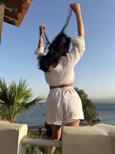 This image, taken in Safaga, Egypt, captures a person from behind, standing outdoors on a sunny day with their back to the viewer. The individual has long, dark hair and is wearing a white, open-knit or crocheted crop top with short sleeves and matching white linen shorts. Both arms are raised above their head, with hands gently grasping and lifting sections of their hair.

The setting is a vibrant coastal landscape. In the background, a large expanse of calm, blue water, likely the Red Sea, extends towards the horizon, where distant, hazy mountains or landmasses are faintly visible. The sky above is clear and bright blue. Lush green palm trees punctuate the scene, suggesting a warm, tropical or subtropical environment.

Architectural details frame the person: a textured white concrete wall or pillar is prominent on the right side, and a wooden slatted structure, possibly part of a railing or pergola, is visible on the left. The bright sunlight indicates it is daytime, and the clear sky suggests fine weather. A notable detail is a light-colored bracelet on the person's left wrist, which appears to have text on it, possibly reading "mamajio" or a similar word. The overall impression is one of leisure and relaxation in a beautiful, sunny resort-like location.