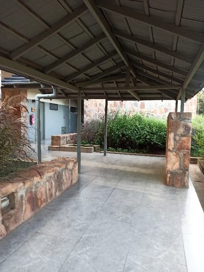 A shaded walkway features a wooden roof structure, open on the sides. Rectangular brick structures line the pathway, with greenery and shrubs visible between them. The tiled walkway appears grey and textured. Further down the walkway, glimpses of a building with gray walls and a door are seen. A small red object hangs on the wall of the building. Tall grass and foliage are at the end of the walkway.
