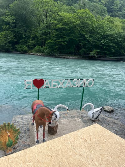 The image displays a scenic view of a turquoise river flowing alongside a lush green forest. In the foreground, there are several decorative objects: a horse sculpture, two swan sculptures, and a peacock sculpture. A sign with Cyrillic lettering, which reads "Я ЛЮБЛЮ АБХАЗИЮ" (I love Abkhazia), is positioned near the riverbank. The text and decorative elements suggest a tourist or recreational area. The overall atmosphere appears peaceful and serene, with the vibrant colors of nature and the decorative elements creating an inviting scene. The lighting suggests it might be daytime, possibly late morning or afternoon, with the sun casting some shadows. The weather appears to be clear and pleasant, conducive to outdoor activities. The setting seems to be a riverside promenade or park, possibly in Abkhazia, given the text on the sign.