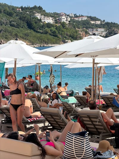 The image depicts a crowded beach scene in Drobnići, Montenegro. The primary subjects are a diverse group of people relaxing on lounge chairs under large white umbrellas. They are dressed in swimwear, and the atmosphere suggests a leisure activity, likely sunbathing and enjoying the beach.
The setting is a beach with clear blue water, indicating good weather and likely a warm time of day, possibly midday or afternoon given the bright sunlight. In the background, a hillside covered in lush green trees and dotted with white buildings, characteristic of Mediterranean architecture, provides a picturesque backdrop. This architectural style and the presence of the Adriatic Sea strongly suggest a coastal location.
Notable details include various beach accessories such as towels, a striped beach bag, and hats. The people are engaged in individual activities such as reading on their phones, conversing, or simply lounging. There are no overt signs of specific events, but the sheer number of people implies a popular tourist spot. The overall mood appears relaxed and peaceful, as one might expect from a beach vacation.
There is some visible text, but it is too blurry and fragmented to be legible. The image is oriented vertically, with the sky and sea at the top, and the beach and people filling the lower portion. The composition emphasizes the density of beachgoers and the vibrant natural surroundings.