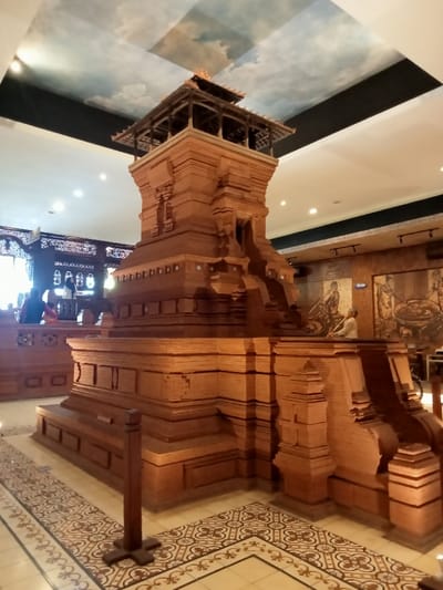 The image shows a wooden model of a traditional Javanese building inside a museum. It is located in the city of Kudus, Indonesia, at coordinates -6.809, 110.843. The model is made of intricately carved wood and features a tiered structure with multiple levels. The building appears to be a miniature replica of a temple or mosque. The model is positioned on a patterned tile floor, suggesting the interior of a museum or exhibition space. The ceiling is decorated with a painted sky, adding to the sense of realism. The model is the only subject in the image, with no people visible. The scene is illuminated by artificial lighting. The overall impression is one of intricate detail and cultural significance. The model is likely a representation of an important religious or historical building.