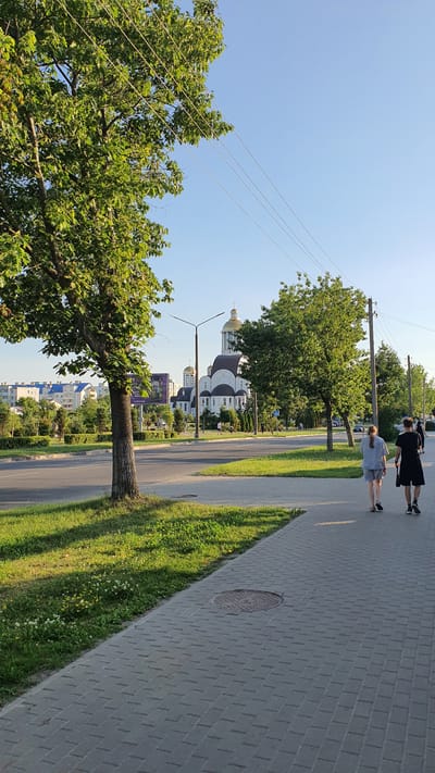 A tree with lush green leaves is in the foreground on the left, casting a shadow on the grassy area next to a paved sidewalk. A road runs parallel to the sidewalk. In the background, a large white church with a golden dome stands prominently. Several trees and some residential buildings are visible behind the church. Two people, a male and a female, are walking away from the camera on the sidewalk. The sky is clear and blue. This image was taken in the city of Salihorsk, Belarus.