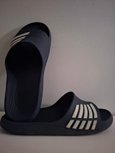 The image shows a pair of dark blue Adidas slide sandals. One sandal is positioned upright, with the other sandal lying flat in front of it. The sandals are made of a textured material and have the signature white Adidas stripes on the upper strap. The background is a plain, textured off-white wall. The foreground is a white surface, likely a shelf or table. There are no people visible in the image. The scene appears to be indoors, possibly in a domestic setting. There is no information or visual evidence within the image to suggest the location is in Huntingdonshire, United Kingdom.