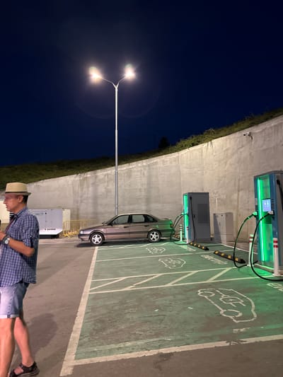 The image depicts an outdoor scene at an electric vehicle (EV) charging station in Kalotina, Bulgaria. The station is marked by green-painted parking spaces with white EV vehicle icons and the word "electric". Two charging units are visible, illuminated with green lights, indicating functionality.

A silver car is parked in one of the charging spots. Adjacent to the car is a white rectangular unit, possibly related to the charging infrastructure.

A man wearing a plaid shirt, denim shorts, sandals, and a straw hat stands at the edge of the frame.

A tall lamppost with two lights illuminates the surrounding area. The backdrop consists of a dark night sky and a concrete structure covered in vegetation.