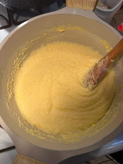 A close-up, overhead shot shows a wooden spoon stirring a pot of creamy, yellow polenta. The polenta is thick and appears to be in the process of cooking, with some bits sticking to the side of the pot. The pot itself is light gray with wooden handles and sits on a stovetop. The background is blurred, but a dark pot and a white bucket are partially visible. The scene is set in a kitchen.