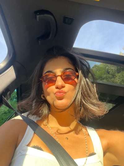 The image presents a close-up, angled portrait of a person, likely a woman, inside a car, bathed in bright, warm sunlight. The subject is positioned in a car seat, with a dark seatbelt visible across their chest. Their dark brown hair, styled with volume, catches the sunlight, showing lighter highlights. They are wearing amber-tinted sunglasses with a tortoise-shell frame, a white top, and multiple delicate gold necklaces, one featuring small, light-colored beads or pearls, and another with a small gold charm. An intricate, dark tattoo, possibly floral or mandala-like, is visible on their upper rib cage, partially concealed by the armpit area. The person has a subtle pout and looks directly towards the viewer, suggesting a posed selfie or photograph.

The interior of the vehicle is visible, including a dark overhead grab handle and a sunroof through which a clear blue sky and hints of green foliage from trees can be seen. The strong, natural light casts pronounced shadows, indicating a sunny daytime setting, possibly during the golden hour. The overall scene conveys a relaxed moment, potentially during travel. Based on the context provided, this moment occurs in Èze, France. No visible text is present in the image.
