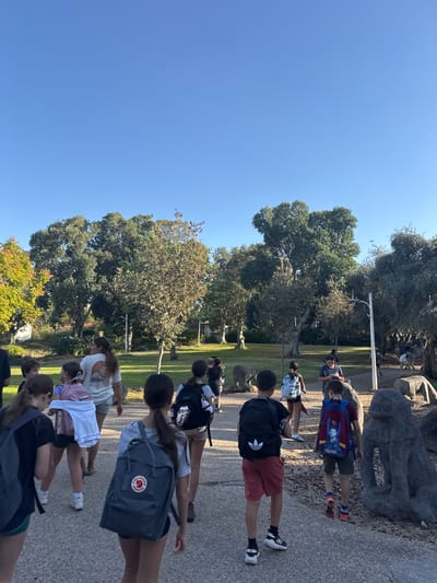 The image displays an outdoor scene in the Gilboa Regional Council, Israel, under a clear, bright blue sky. A group of approximately 10-15 individuals, primarily children and adolescents, are walking away from the camera along a light-colored, pebbled paved path that extends into a park-like environment. Most individuals are wearing casual clothing and backpacks of various colors and brands.
In the mid-foreground, a female with long dark hair wears a gray t-shirt and a gray Fjallraven Kanken backpack. To her right, a male child wears a dark t-shirt, red shorts, and a black Adidas backpack. Further to the right and slightly ahead, another male child wears a blue and red themed backpack. Several other individuals are visible further along the path and on the surrounding grass.
The path is bordered by green grass and numerous trees with varied green foliage, forming a dense natural backdrop. To the right of the path, and continuing into the background, are multiple stylized, rough-textured grey stone sculptures resembling animals, possibly large cats or primates. A light pole is visible in the middle-right background. The scene is illuminated by bright natural daylight.
