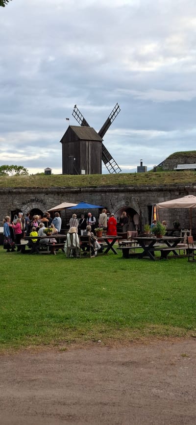 The image depicts an outdoor gathering in front of a stone building with arched entrances, which appears to be a historical structure in Kuressaare, Estonia. Above the stone building, there is a grassy embankment topped with a wooden windmill with visible blades. A group of people are congregated near the entrances, some standing, others seated at picnic tables set up on a lawn. Several umbrellas provide shade. The overall atmosphere suggests a casual social event or a tourist attraction. The sky is overcast.