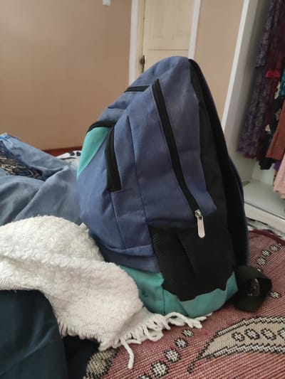 The image depicts a backpack resting on a red patterned rug. The backpack is primarily blue with teal and black accents, featuring zippered compartments. To the left of the backpack, a white textured cloth is draped over dark blue fabric, which is itself on top of a blue patterned textile. In the background, a wall of a neutral color is visible, along with a closet door and clothing items hanging inside a white closet. The scene appears to be indoors, possibly in a bedroom or dressing area, in the city of Kipchak, Tajikistan. There are no people or animals present in the image.