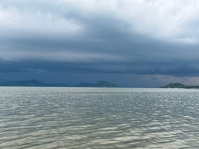 The image depicts a wide expanse of calm water under a dramatic, overcast sky. Dark, heavy clouds dominate the upper portion of the frame, with lighter clouds near the horizon. In the distance, low-lying islands or peninsulas with green vegetation are visible. The water's surface is rippled, reflecting the subdued light and creating a textured pattern. There are no visible people or specific structures that clearly identify the location within Ban Bang Khao, Thailand, beyond the general coastal environment. The overall impression is one of a serene, possibly pre-storm seascape.