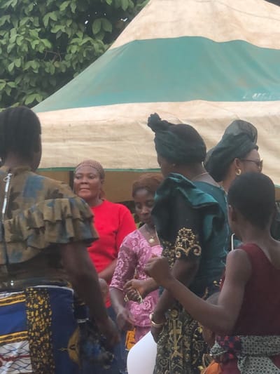 This image captures an outdoor gathering in Awka Etiti, Nigeria. The scene is set against a backdrop featuring a large tent with a white body and a teal green horizontal stripe near its apex, with green tree foliage visible above and to the left of the tent.

In the foreground, a person with dark skin is seen from the back on the left, wearing a brown and blue patterned top with ruffled sleeves, and a blue and yellow patterned skirt. To their right, another person, also with dark skin, is visible from the back wearing a dark red sleeveless top and a patterned waist wrap.

In the midground, several individuals, primarily women, are present. A woman with dark skin and a brown headwrap, wearing a red short-sleeved top, is centrally positioned and looks slightly to her right. To her left and slightly behind, a woman with light brown hair, wearing a pink lace top and a necklace, looks downwards. To the right of the central red-shirted woman, another woman with dark skin, wearing a dark green headwrap and a dark green garment featuring intricate gold beading on the sleeve/shoulder, looks downwards. Partially visible on the far right is a woman with a dark green headwrap, glasses, and a gold hoop earring.

The individuals are standing in close proximity, suggesting a social event. No overt actions or interactions are clearly discernible beyond their presence at the gathering.
