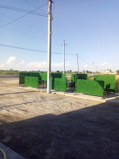 The image shows an outdoor seating area with several small tables and chairs, enclosed by artificial green hedges. A concrete pole with electrical wires runs through the scene. In the background, there is a road and some buildings, suggesting an urban or semi-urban environment. The ground is covered with gravel and dirt. The sky is clear and blue with a few scattered clouds. The overall impression is one of a simple outdoor dining or relaxation space.