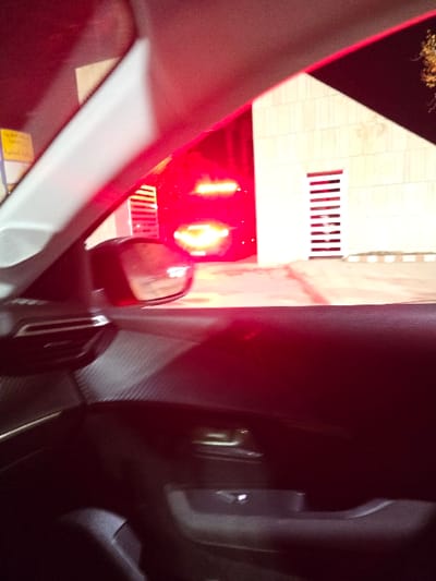 The image shows an interior car view at night. Through the car window, a building is visible, illuminated by red and yellow lights, potentially emergency lights. The car's side mirror reflects the building's lights. The car's interior details include the dashboard, air vent, and various interior components, all shrouded in darkness except where the red lights cast a dim glow.