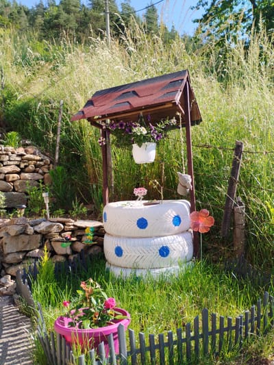 This image depicts a whimsical garden scene in the city of Smilyan, Bulgaria. The central focus is a decorative well structure constructed from wooden posts and a shingled roof. Suspended from the structure are white flower pots overflowing with purple and white petunias. The "well" itself is formed by a stack of three whitewashed tires, each adorned with blue polka dots.

To the left of the well, a meticulously crafted stone wall rises, with smaller rocks and plants nestled within its crevices. A solar-powered garden light is visible near the base of the wall. Scattered amongst the grass are small, colorful garden ornaments, including a ladybug, a frog, and a butterfly-shaped decoration.

In the foreground, a vibrant pink planter holds a blooming rose bush with bright pink flowers. A small, dark grey picket fence encloses this planter. To the right of the well, a thin wire fence with wooden posts runs horizontally. A translucent orange flower stake is positioned in front of the tires. A grey, bird-like ornament hangs from a string near the well structure.

The environment is lush and overgrown, with tall grasses and various green plants dominating the background. Sunlight illuminates the scene, casting shadows and highlighting the textures of the stone wall, tires, and foliage. The sky above is a clear, bright blue. The overall impression is one of a creatively decorated and vibrant outdoor space.