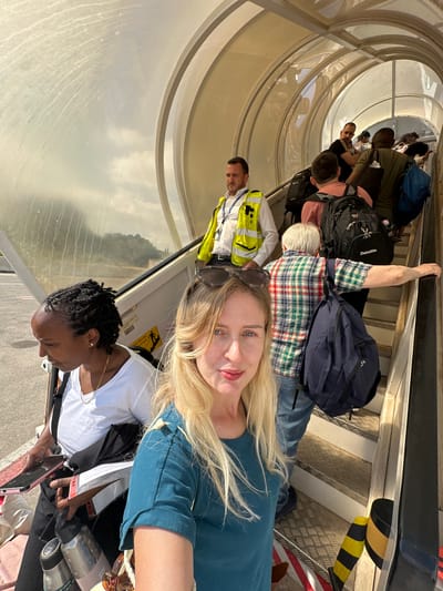 In Yehud-Monosson, Israel, people are boarding a plane via an outdoor staircase. The image features a woman in the foreground taking a selfie, with another woman walking up the stairs to her left. A man in a yellow vest is visible further up the stairs, seemingly directing passengers. The environment appears to be an airport tarmac under a cloudy sky, with a covered walkway leading up to the aircraft. Passengers are carrying luggage, including backpacks.