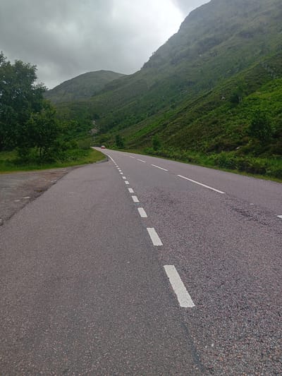 The image shows a road in a mountainous, green landscape. The road, made of asphalt, has white dashed lines painted on its surface. On the left side of the road, a steep, grassy hillside rises up, covered in lush green vegetation. In the distance, the outline of a mountain is visible against a cloudy sky. A few trees are scattered along the roadside, with one prominent dark green tree in the foreground. The sky is overcast, suggesting a cloudy or rainy day, which is typical of the United Kingdom. There is a small, red object, possibly a car, visible in the distance on the left side of the road. The image appears to be taken from a low angle, looking down the road as it curves to the left. The overall scene evokes a sense of travel and exploration through a natural, rugged landscape. The location is identified as Invergarry, United Kingdom. There is no visible text in the image.